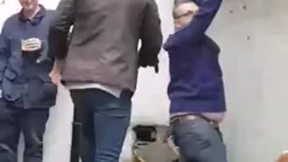 Man in brown jacket tries to help bald friend in blue climb wall fails to only lift one leg